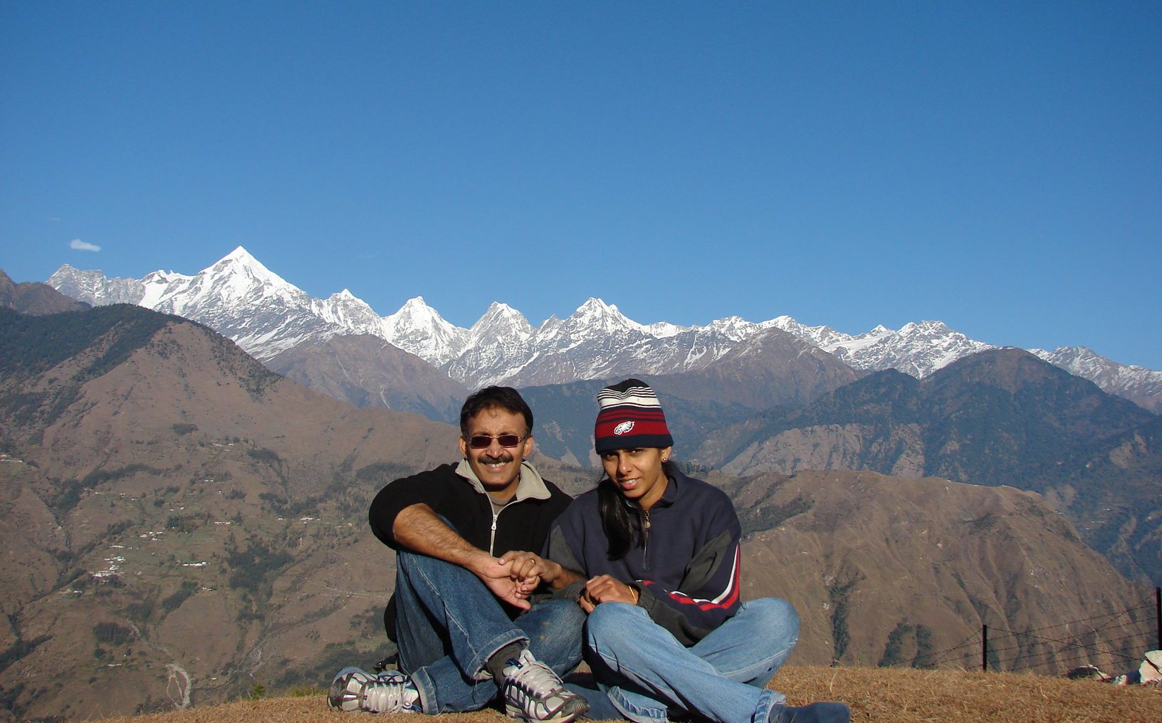 A couple squatting on the ground with mountains and snowcapped peaks behind under a blue sky