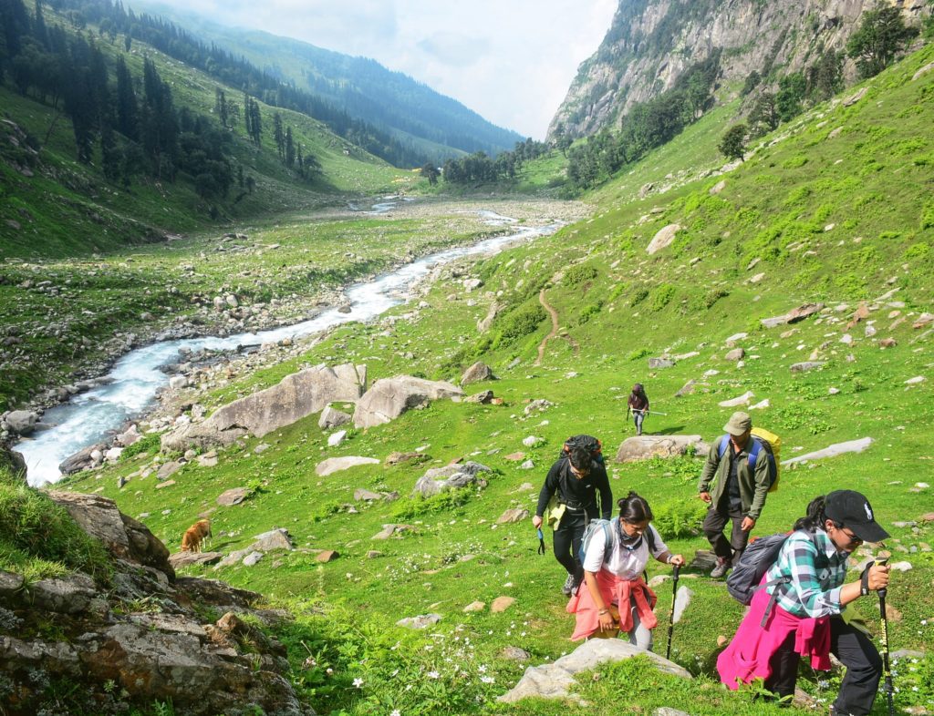 Trekkers climbing up in a green meadow with a river flowing under a blue sky