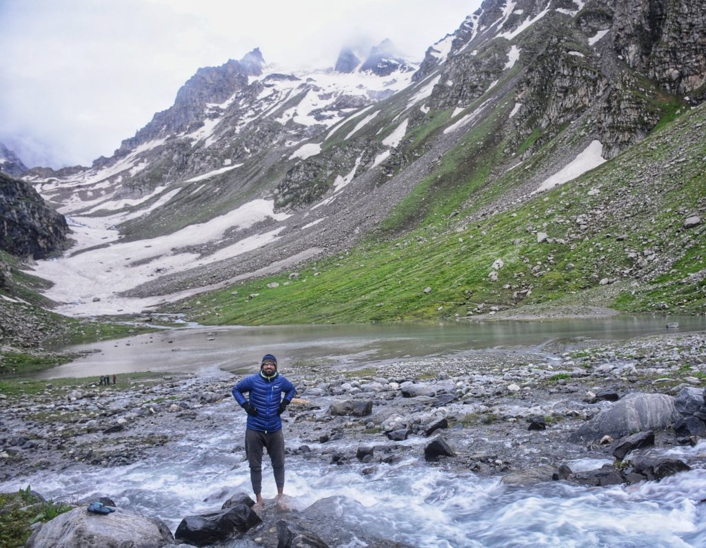 A mountain valley with a snow path in the background and a flowing river and a trekker standing