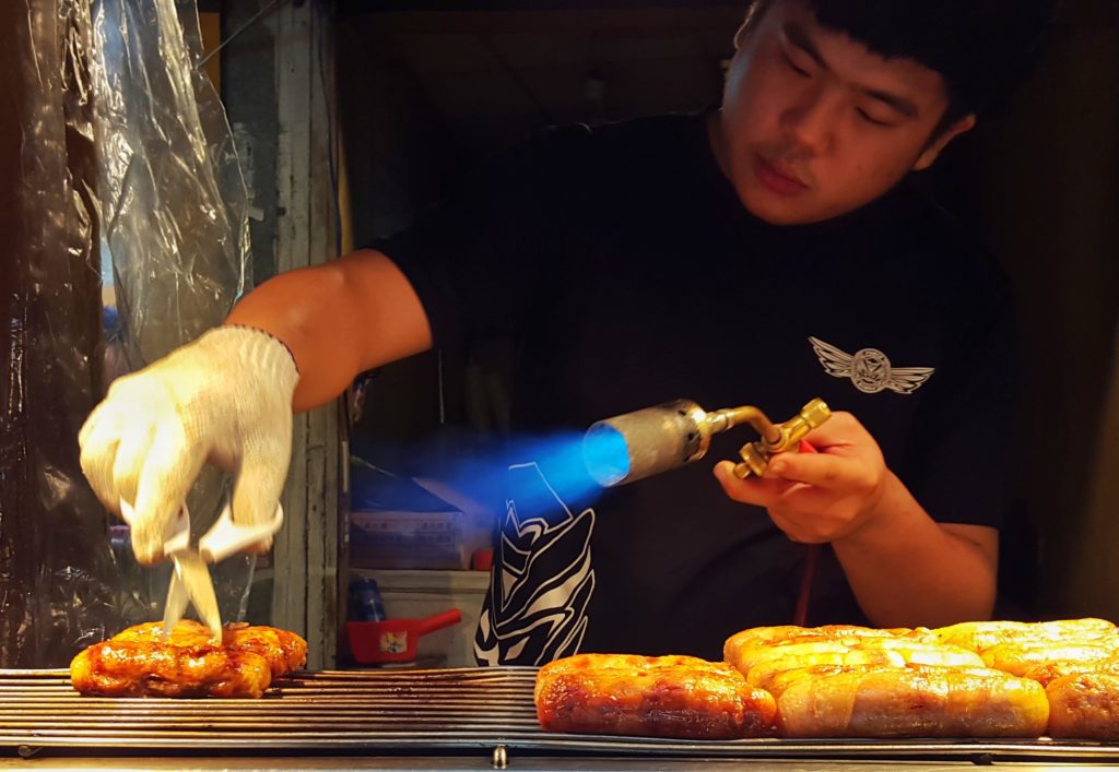 A street food vendor using a blow torch and scissors to cook a piece of meat.