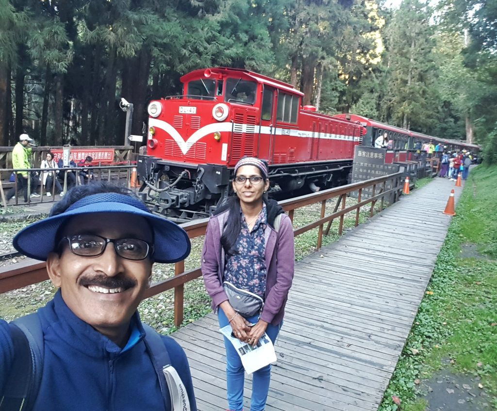 A couple posing in front of a train at a station by the woods.