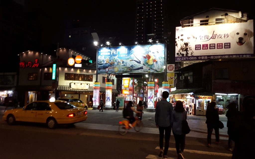 City street at night with neon lit shop fronts and people and cars