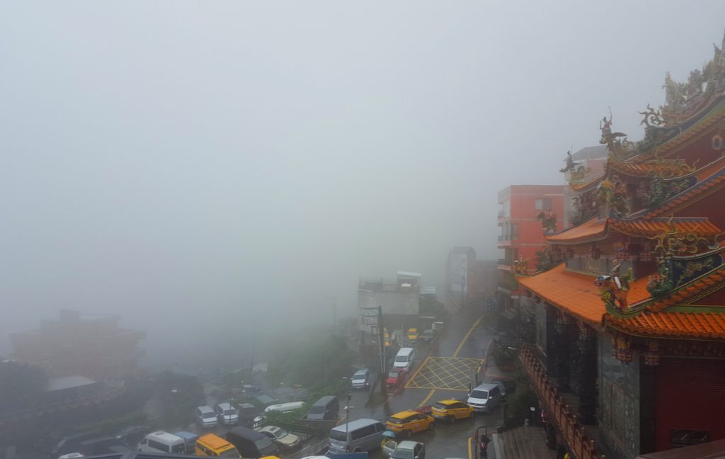 Scene of a mountain town with a temple on a street filled with cars during a foggy day