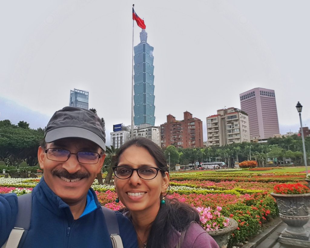 A couple posing at a park overlooking tall buildings and a skyscraper with a flag furling