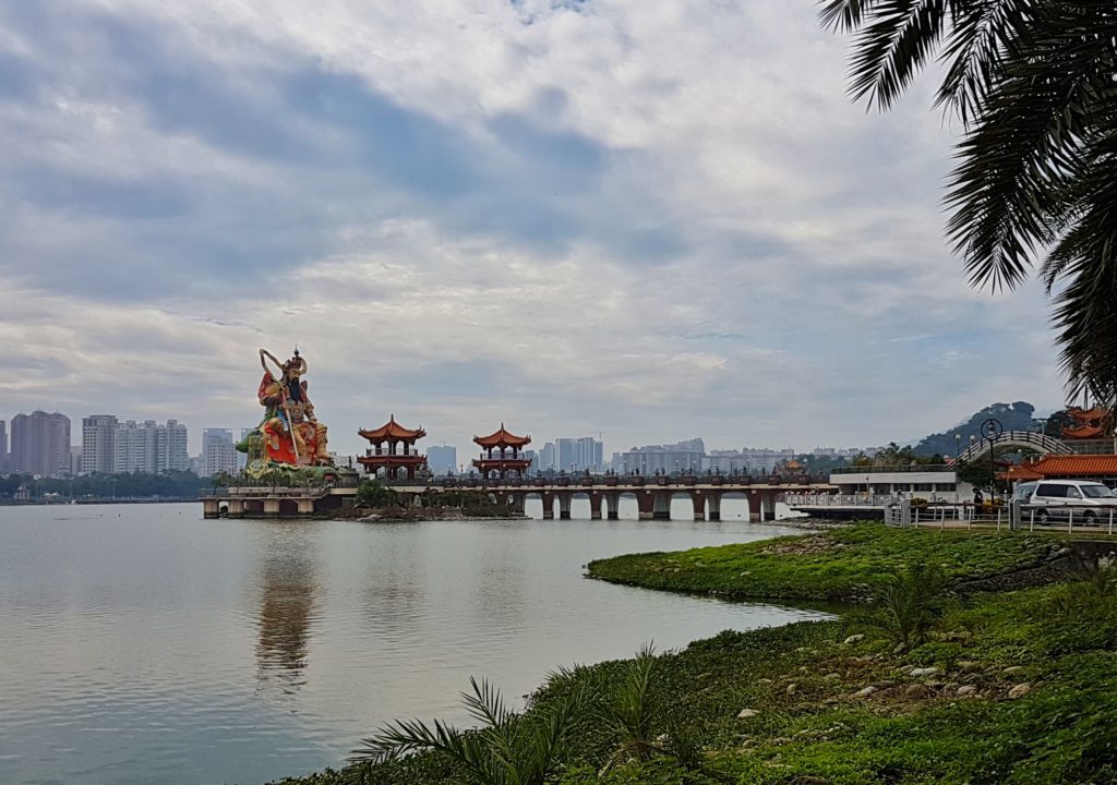 A lake with green banks and a big statue and pagodas in the middle and buildings in the background