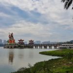 A lake with green banks and a big statue and pagodas in the middle and buildings in the background