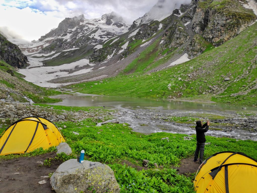Snow clad mountain, green meadows along a flowing river and yellow tents pitched with a man taking a photo