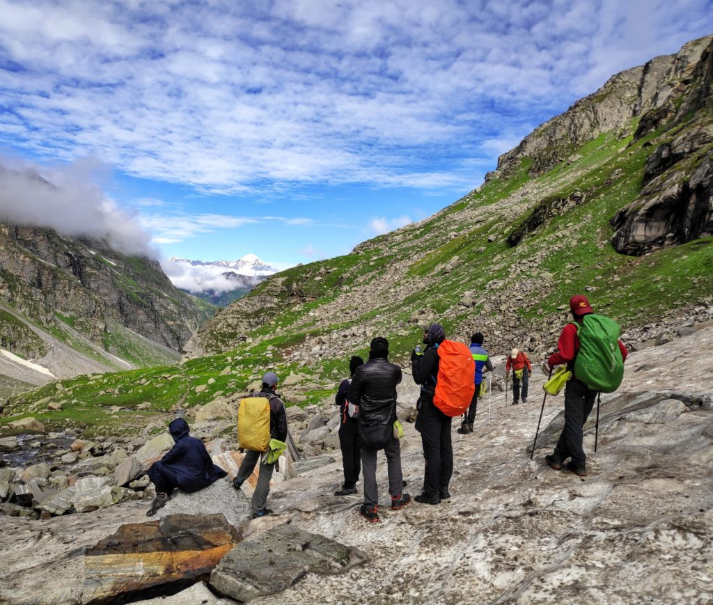 Trekkers standing on a snow clad mountainside looking at a green meadow and snow capped mountain peak in the distance