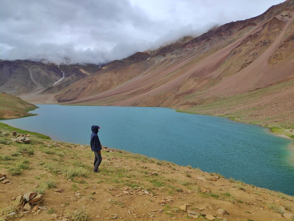 A man stands at the edge of a mountain lake covered with barren hills