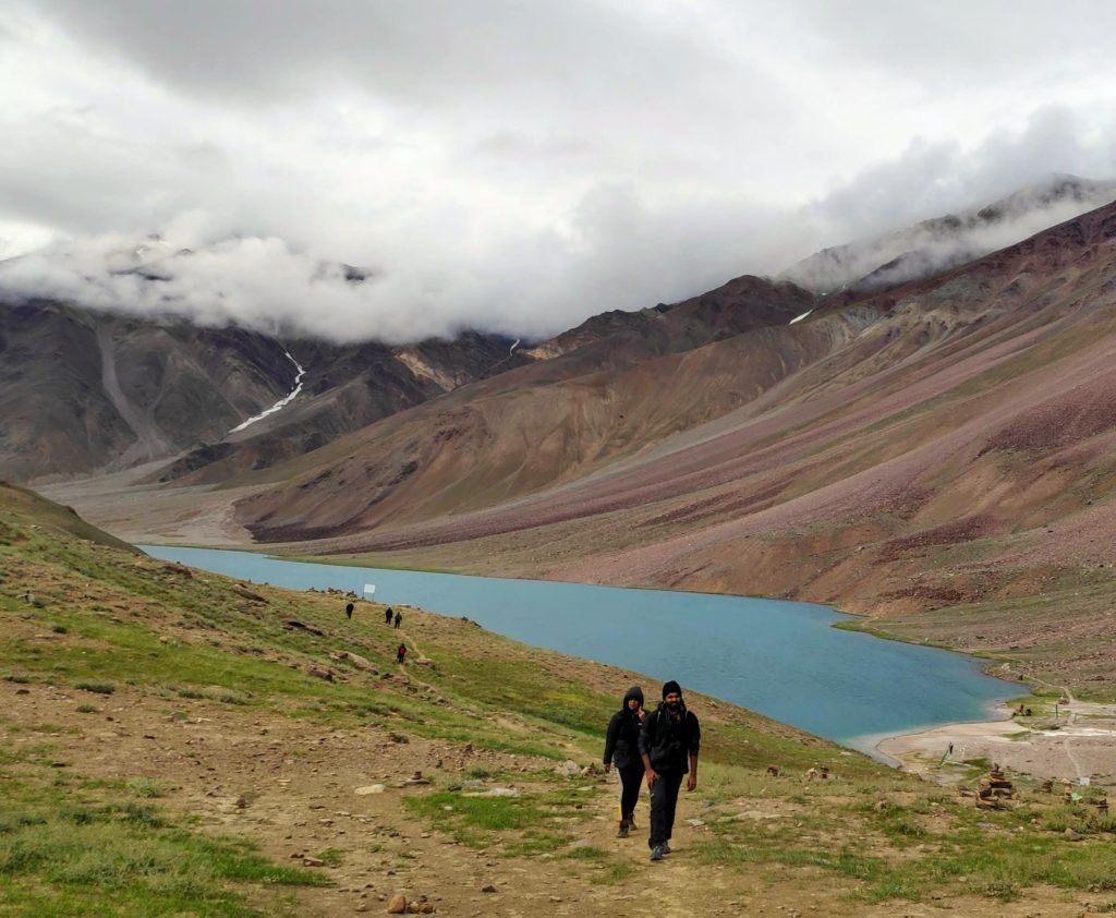 Two travellers walking near a mountain lake with turquoise water and surrounded by grey barren hillside under a cloudy sky